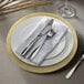A white Charge It by Jay glass charger plate with a gold burst design and silverware on a white napkin.
