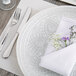 A white Charge It by Jay glass charger plate with silver trim on a table with a napkin and flowers.