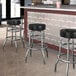 Three Lancaster Table & Seating Black Vinyl Double Ring Swivel Bar Stools in front of a counter in a bar.