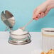 A hand using a spoon to scoop sugar from a Tablecraft glass condiment jar.