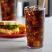 A close-up of a clear plastic GET tall tumbler filled with soda and ice next to a sandwich.