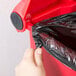A hand pulling a black bag out of a red Rubbermaid commercial trash can.
