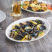 A Tuxton AlumaTux china platter with mussels in white wine sauce and a lemon wedge on a table with a glass of wine.