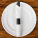 A white plate with a black self-adhering paper napkin band wrapped around a napkin with a fork and knife inside.