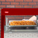 A person's hand holding a tray of french fries in a Hatco red stainless steel countertop food warmer.