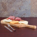 An American Metalcraft olive wood serving board with meat and cheese on it next to a fork and knife.