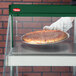 A hand in glove holding a pizza in a Hatco countertop glass display case.