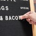A person using Aarco Helvetica Universal Single Tab Letter and Number Set to point to a sign that says "Bacon" on a counter.