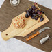 A Franmara stainless steel cheese knife with a bamboo handle on a wooden cutting board with grapes.