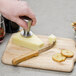 A hand using a Franmara stainless steel cheese knife with a bamboo handle to cut cheese on a counter.