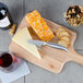 A Franmara stainless steel hard cheese and chocolate knife on a cutting board with cheese and crackers next to a bottle of wine.