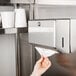 A hand pulling a paper towel from a Bobrick stainless steel paper towel dispenser on a wall.