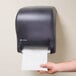 A hand holding a paper towel in a black San Jamar Tear-N-Dry Essence hands free paper towel dispenser.