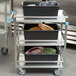 A Lakeside stainless steel utility cart with three shelves holding black containers with plates on them.
