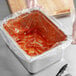 A person using a 1/2 size deep steam table pan liner to hold a plastic bag with red sauce inside.