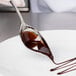 A hand holding a Mercer Culinary Saucier Spoon with chocolate syrup over a plate.