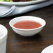 A white porcelain sauce dish filled with red sauce on a table.