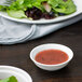 A white porcelain sauce dish filled with red sauce next to a plate of salad.