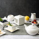 A white square teapot with a lid on a table set with white dishes and fruit.