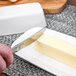 A person's hand using a knife to cut butter on a rectangular white plate.