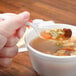 A hand holding a Dart white plastic teaspoon over a bowl of soup.