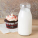 A cupcake with frosting on top and a glass of milk on a wooden table. A close up of an American Metalcraft glass bottle of milk.