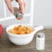 A hand adding salt to a bowl of pasta from a Tablecraft Nostalgia glass salt shaker.