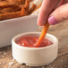 A person dipping a carrot stick into a Carlisle ivory ramekin of ketchup.