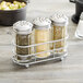 A Tablecraft glass spice shaker in a metal rack filled with spices.