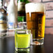An Arcoroc square shot glass with yellow liquid next to a glass of green liquid.
