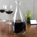An Arcoroc square glass carafe filled with red wine on a table.