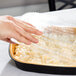 A hand using a plastic container to put food in a black and gold aluminum foil pan with a dome lid.