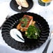 A Fineline black plastic plate with a piece of salmon and a glass of wine on a table.