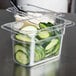 A clear plastic food pan with cucumbers and cheese on a counter.