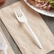 A white plastic Choice medium weight fork on a napkin next to food on a plate.