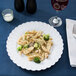A Fineline Flairware white plastic plate with pasta and broccoli next to a glass of wine.