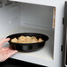 A hand holding a Pactiv Newspring VERSAtainer bowl of food in a microwave.