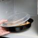 A hand holding a Pactiv Newspring VERSAtainer oval plastic container with a plastic lid over food in a microwave.