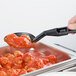 A person holding a Cambro black salad bar spoon over a pan of food.