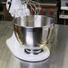 A white KitchenAid stand mixer with a whisk in a white stainless steel mixing bowl with a handle.