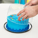 A hand reaching for a blue cake in a plastic display container with a clear dome lid.