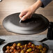 A hand holding a Lodge cast iron skillet cover over food cooking in a skillet.