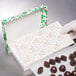 A hand in a glove opening a white 3-ply glassine candy box pad with a gold floral pattern to reveal chocolates.