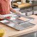 A person wearing gloves is holding a rectangular metal frame above a large metal baking sheet on a wooden countertop.