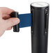 A hand holding a blue retractable belt attached to a black Aarco crowd control stanchion.