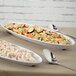 Two white Siciliano oval platters filled with food on a table with silver spoons.