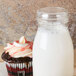 A wooden table with a glass jar of milk and a cupcake with an American Metalcraft round PET milk bottle cover on top.