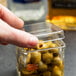 A hand putting a yellow American Metalcraft lid on a small glass jar of olives.