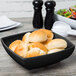 A black Milano square bowl filled with bread rolls on a table.