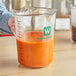 A person using a WebstaurantStore clear plastic measuring cup to pour orange liquid.